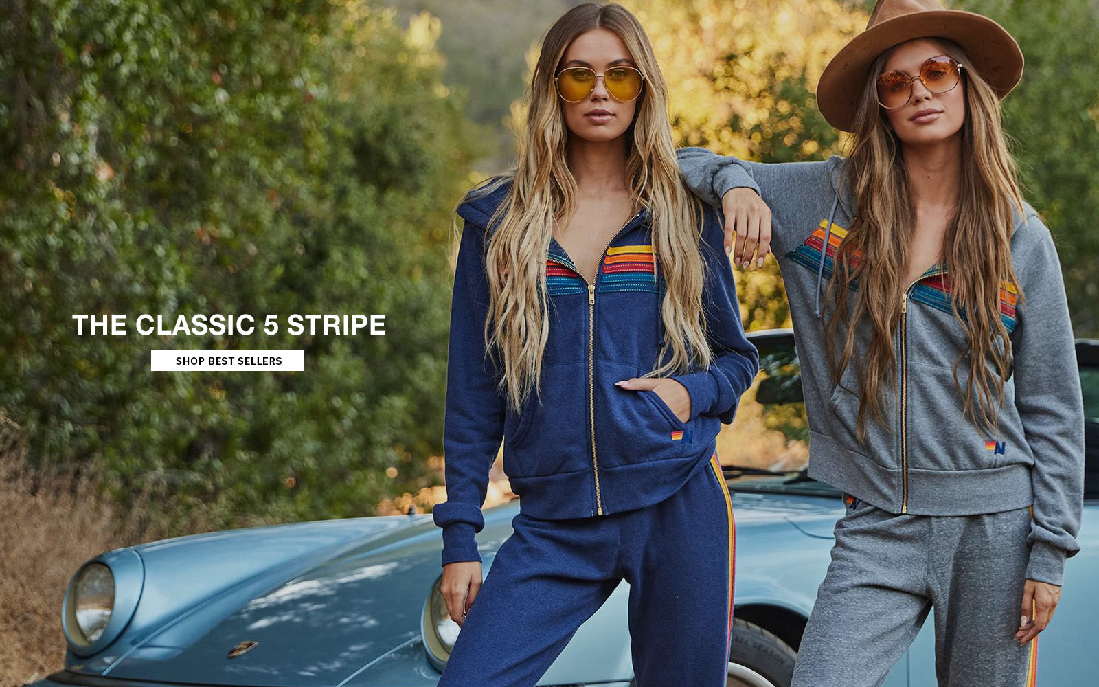 Two women in matching tracksuits standing next to a vintage car with trees in the background.