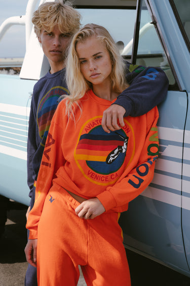 A woman in a bright orange Venice Carnival-themed sweatshirt and matching sweatpants stands in front of a vintage light blue truck, with a man in a dark navy sweatshirt leaning behind her.