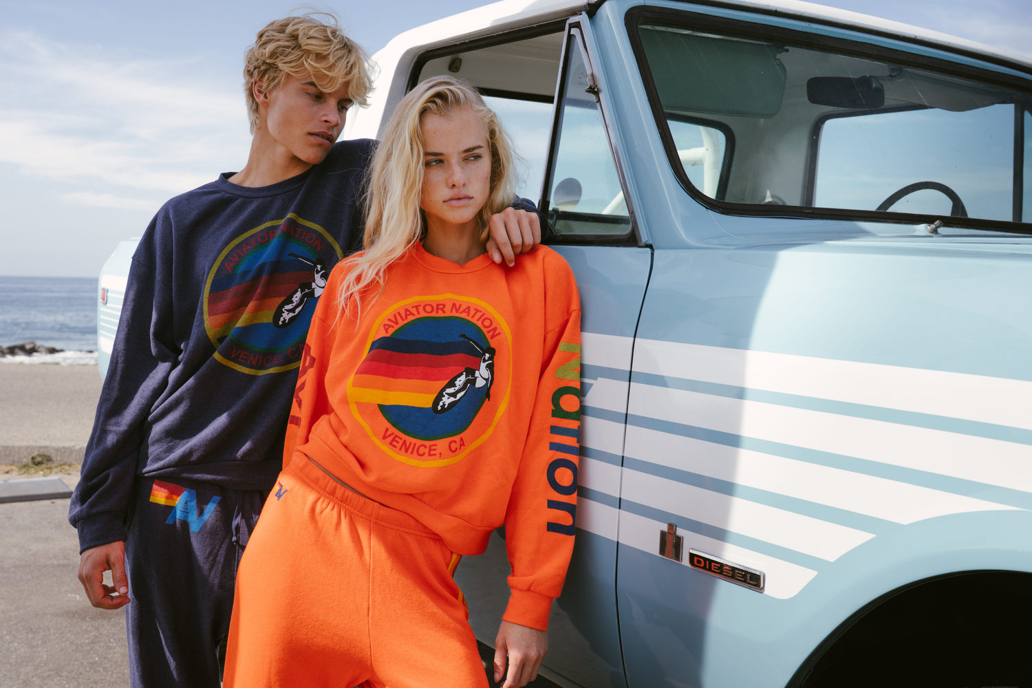 Two models posing next to a vintage light blue pickup truck outdoors. The blonde model in the foreground wears a bright orange crewneck sweatshirt and matching sweatpants with a colorful circular graphic on the chest.