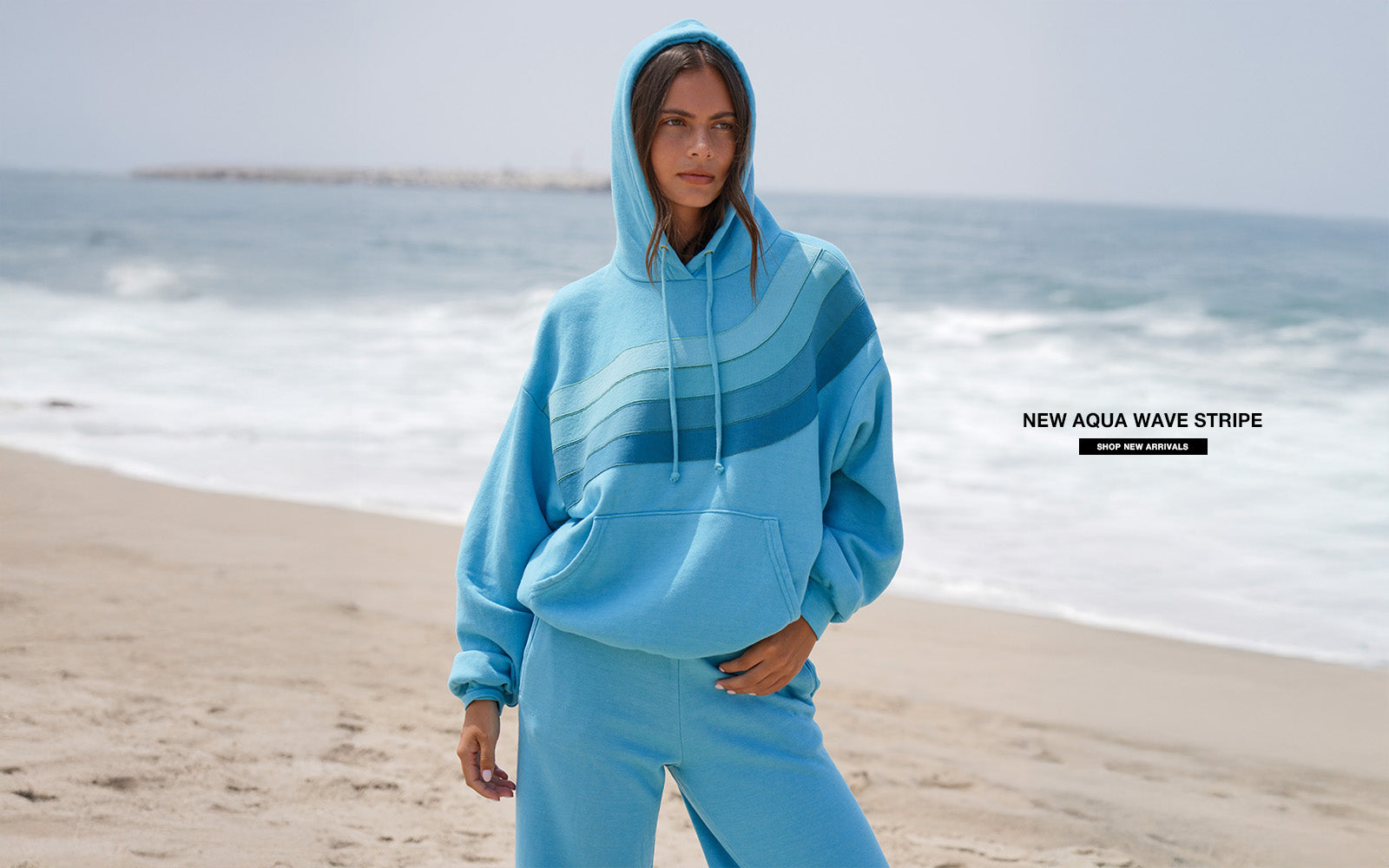 Person wearing a blue hoodie and pants on a beach with ocean waves in the background