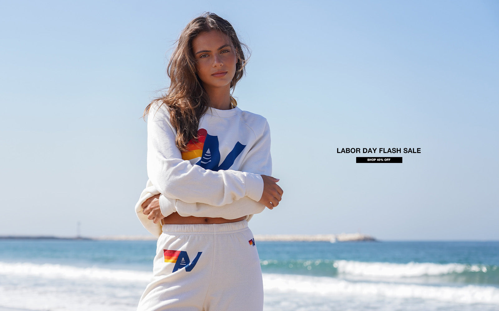 Woman wearing a white sweatshirt with colorful designs on a beach with a Labor Day flash sale banner.