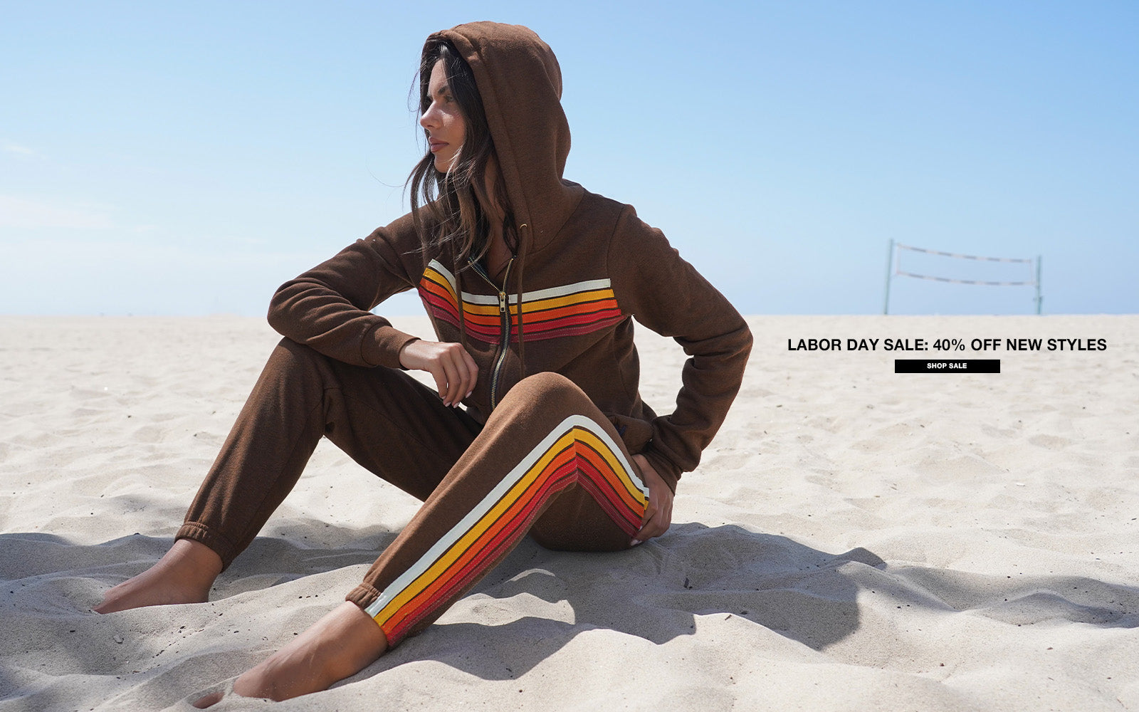 Person wearing a brown hoodie and pants with colorful stripes sitting on a sandy beach.