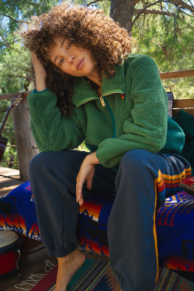 A woman with curly brown hair sits on a wooden deck chair outdoors, wearing a green fleece zip-up jacket and dark blue sweatpants with colorful striped accents.