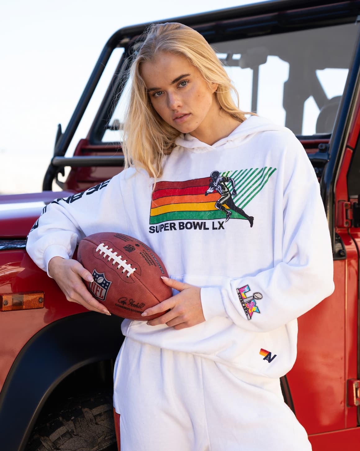 Person wearing a white hoodie with colorful graphics and text, holding a football in front of a red vehicle.