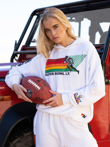Person wearing a white hoodie with colorful graphics and text, holding a football in front of a red vehicle.