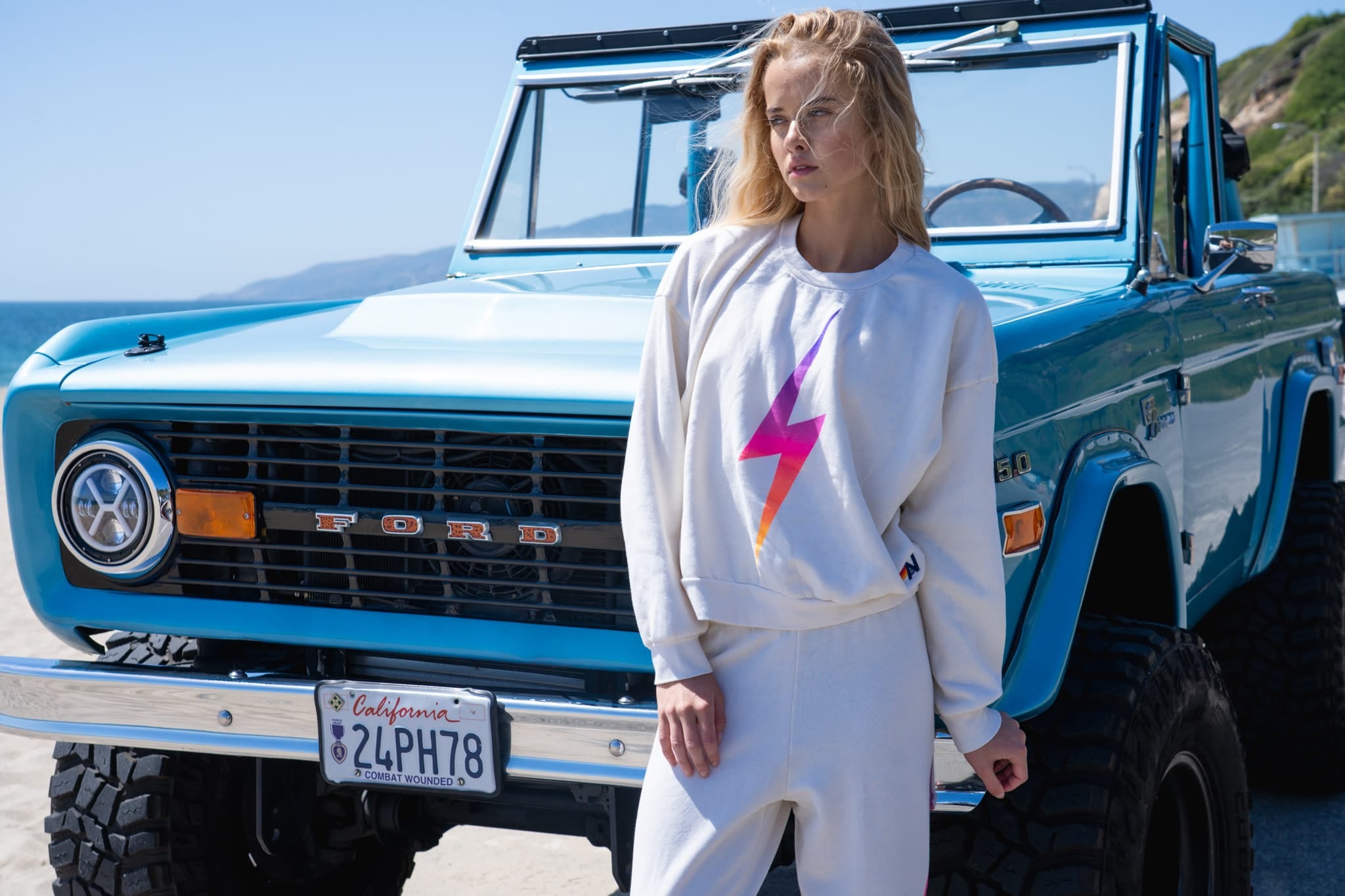 Person wearing a sweatshirt with a colorful lightning bolt design in front of a blue Ford Bronco.