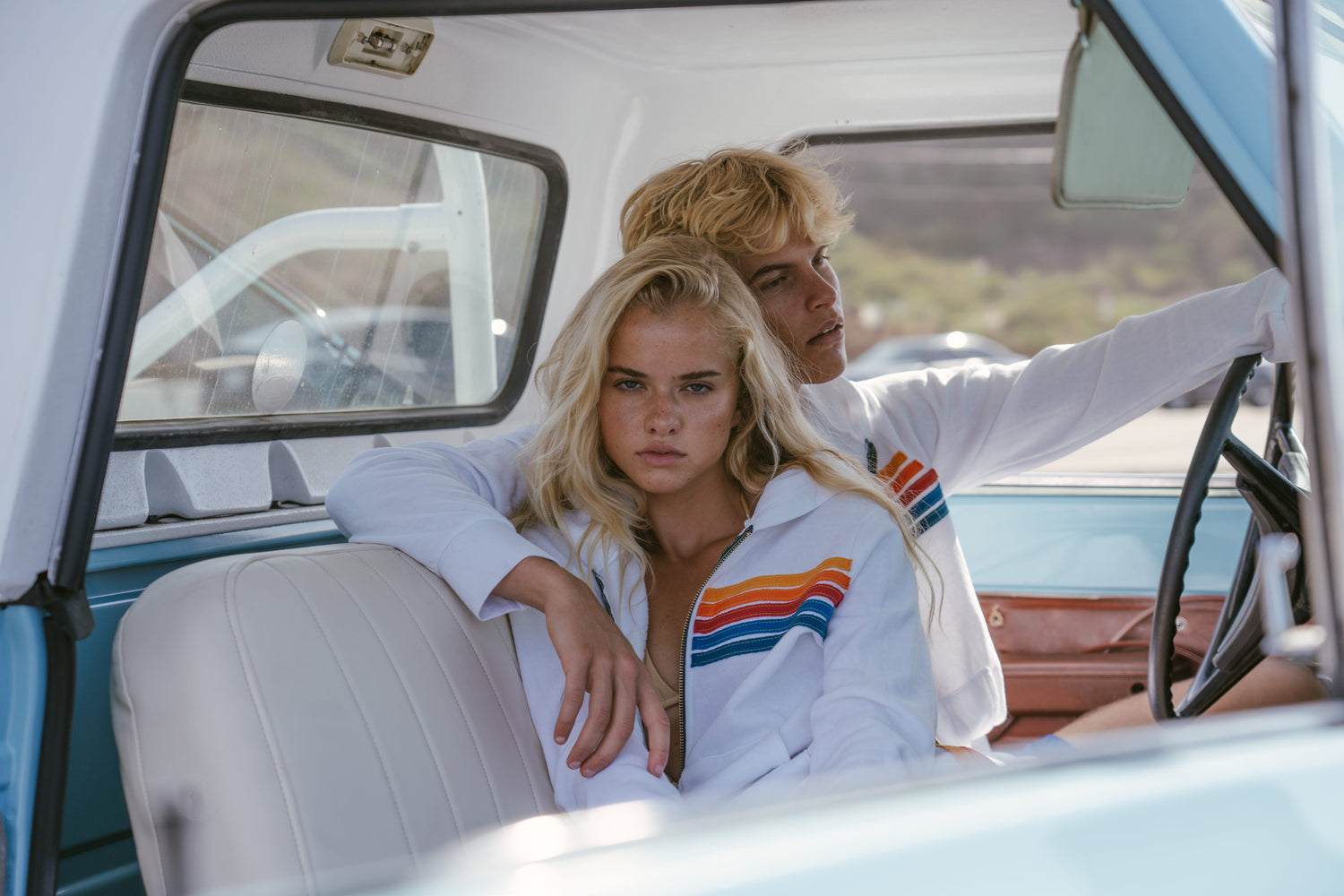 Two models wearing matching white vintage-style striped jackets with orange, yellow, and blue stripes, posed inside a light blue and white classic truck.