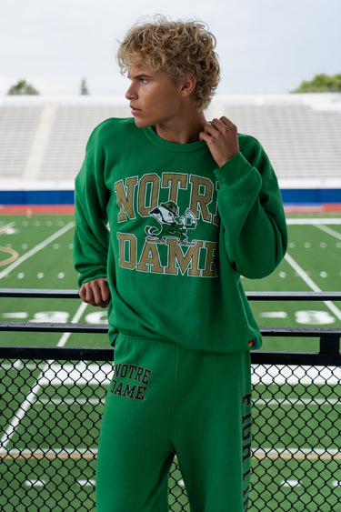 A woman with long blonde hair stands next to a bright yellow classic car outdoors, wearing a matching navy blue zip-up hoodie and sweatpants set with retro rainbow stripes across the chest.