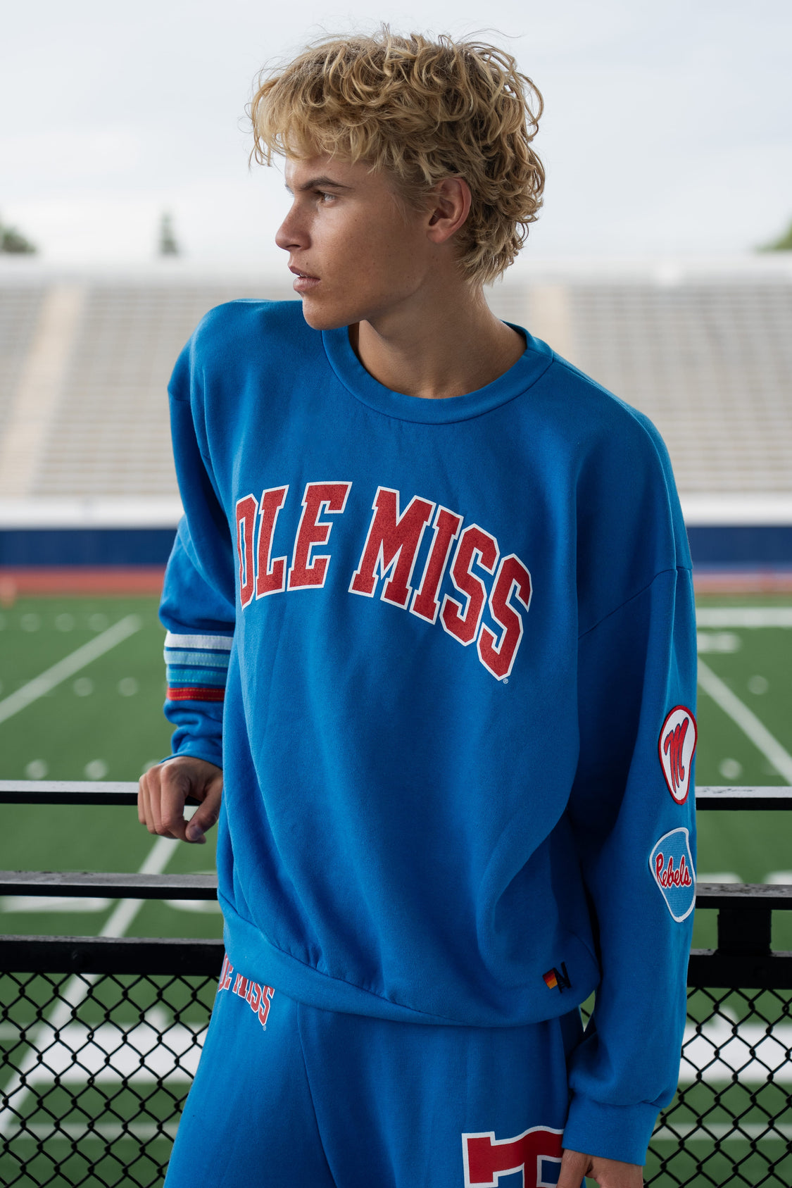 A young man with curly blond hair wearing a bright blue Ole Miss sweatshirt and matching sweatpants, standing on an outdoor athletic field with stadium seating in the background.