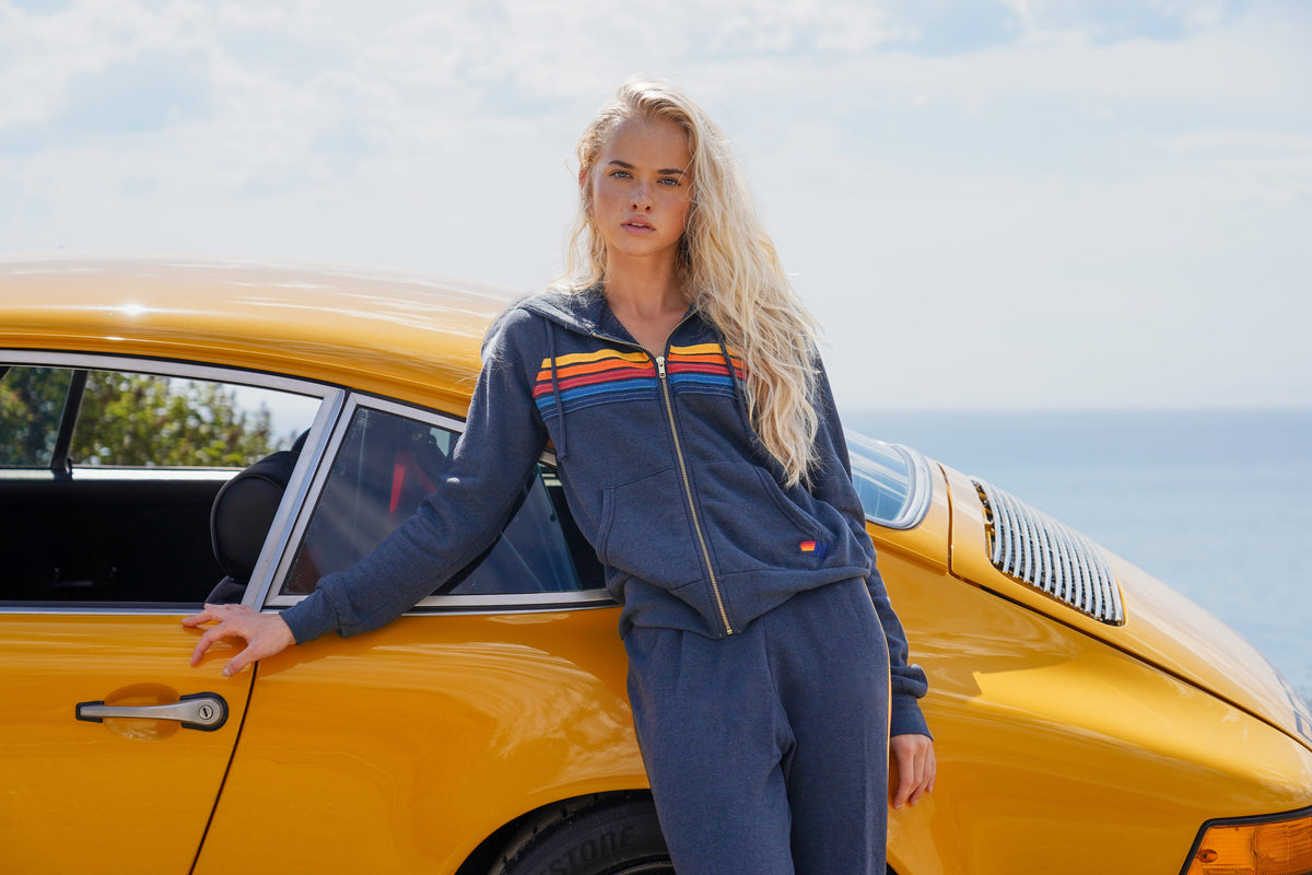 A woman with long blonde hair stands next to a bright yellow classic car outdoors, wearing a matching navy blue zip-up hoodie and sweatpants set with retro rainbow stripes across the chest.