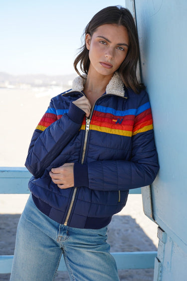 A woman wearing a navy blue puffer jacket with colorful horizontal stripes in red, yellow, and blue across the chest, a sherpa collar, and a zippered front, leaning against a light blue structure outdoors.