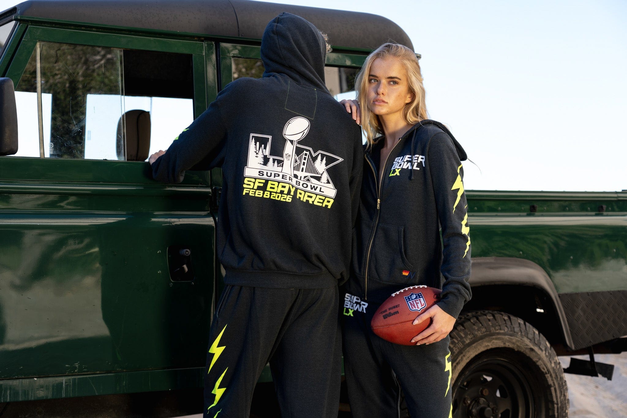 Two people wearing branded clothing standing next to a green vehicle.