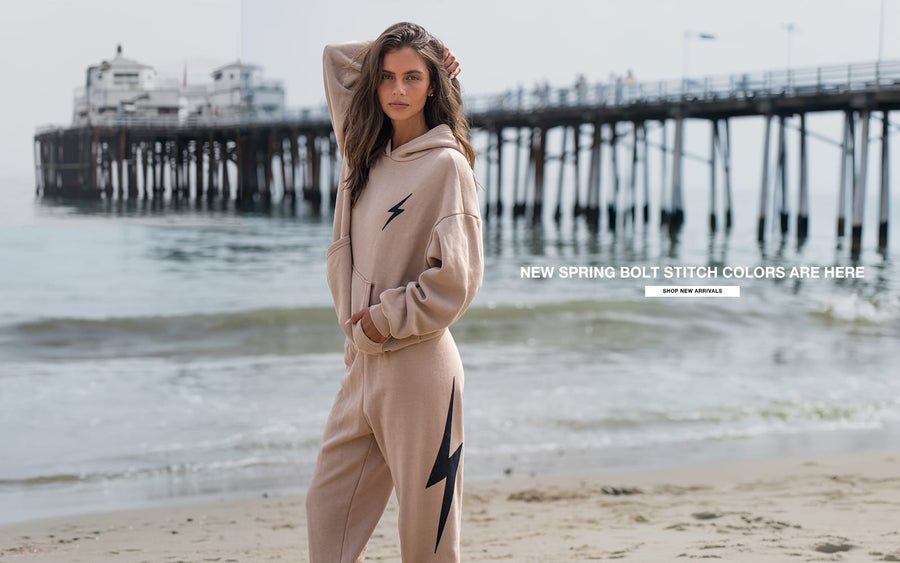 Woman in beige hoodie and pants with black lightning bolt design on beach, with text overlay about new spring bolt stitch colors.