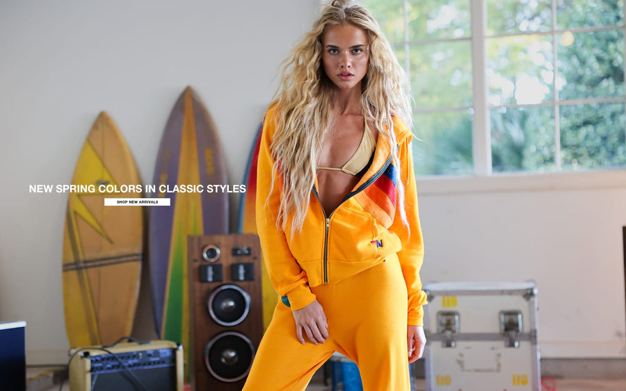 Woman in a bright yellow tracksuit standing in a room with surfboards and a window in the background.