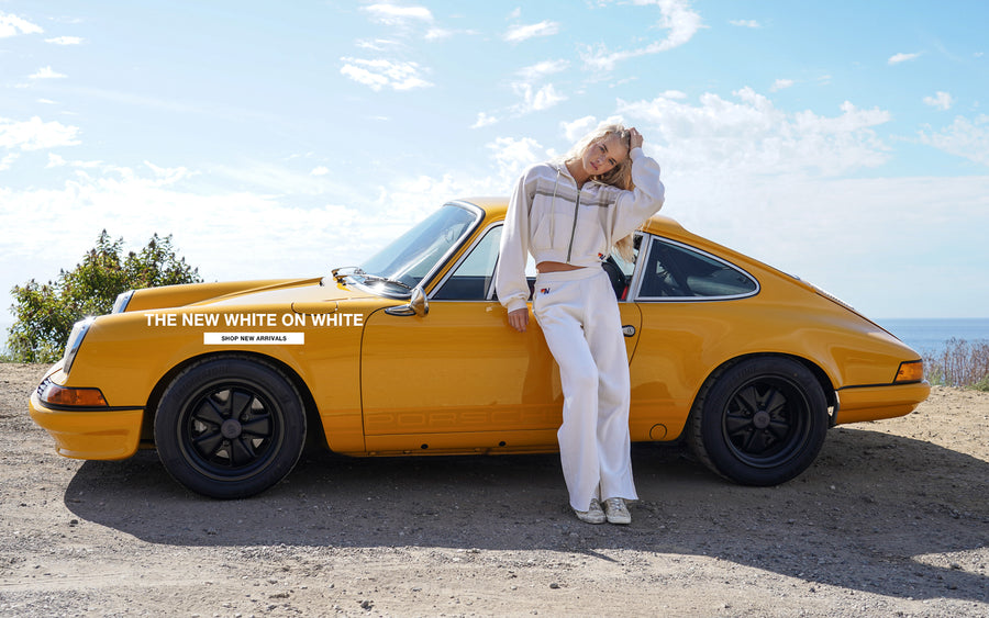 Woman leaning against a yellow Porsche with 'The New White on White' logo on a scenic background
